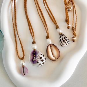 May include: Five necklaces with brown cords and shell pendants. The pendants are a variety of shapes and colors, including a purple and white shell, a brown shell, and a white and black shell.