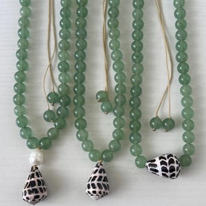 May include: Three necklaces with green beads and seashell pendants. One necklace has a white pearl and a black and white seashell pendant. The other two necklaces have black and white seashell pendants. The necklaces are hanging on a white surface.