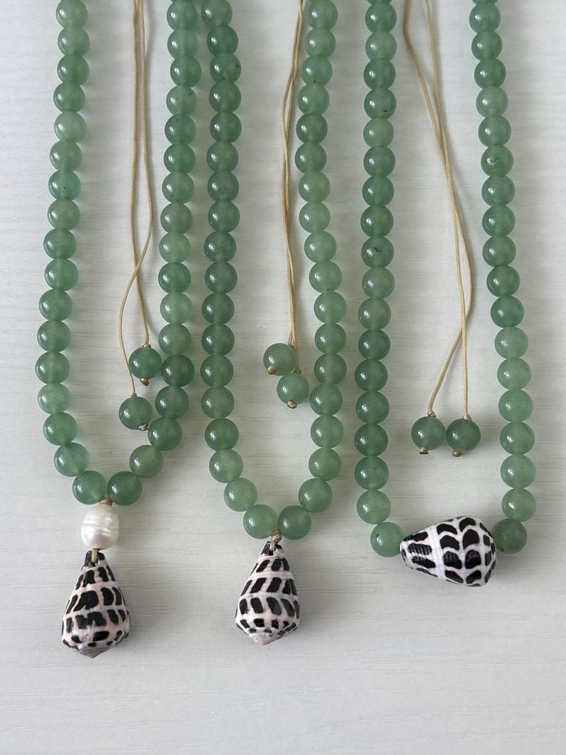 May include: Three necklaces with green beads and seashell pendants. One necklace has a white pearl and a black and white seashell pendant. The other two necklaces have black and white seashell pendants. The necklaces are hanging on a white surface.