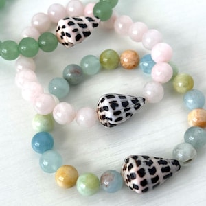 May include: Three beaded bracelets with a beach theme. The bracelets feature a variety of pastel-colored beads, including pink, green, blue, and yellow. Each bracelet has a seashell accent with black and white spots. The bracelets are displayed on a white surface.