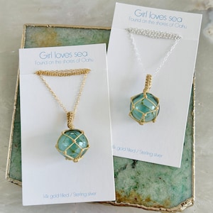 May include: Two necklaces with a glass globe pendant. The globes are encased in a wire cage. The necklaces are on white cards with the text "Girl loves sea" and "Found on the shores of Oahu". One necklace has a gold chain and the other has a silver chain. The card with the gold chain necklace says "14k gold filled/Sterling silver". The card with the silver chain necklace says "14k gold filled/Sterling silver".