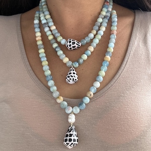 Op de afbeelding: Drie-rijige ketting met aquamarijnkleurige kralen en schelpenhangers. De kettingen hebben een mix van lichtblauwe, crèmekleurige en bruine kralen. De schelpenhangers zijn zwart en wit met een rasterpatroon.