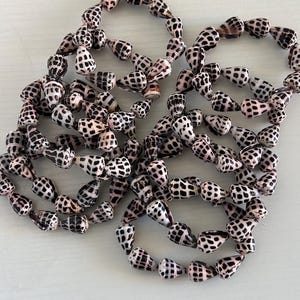 Peut inclure: Gros plan de plusieurs brins de coquillages, chacun avec un motif distinctif de taches noires sur un fond blanc et rose. Les coquillages sont enfil&eacute;s ensemble, formant des formes circulaires. L'image est prise d'en haut.