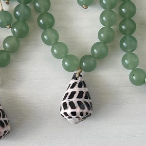 May include: A necklace featuring light green, round beads and a teardrop-shaped pendant. The pendant is a seashell with a black and white pattern. The necklace is strung on a thin, gold-colored cord.