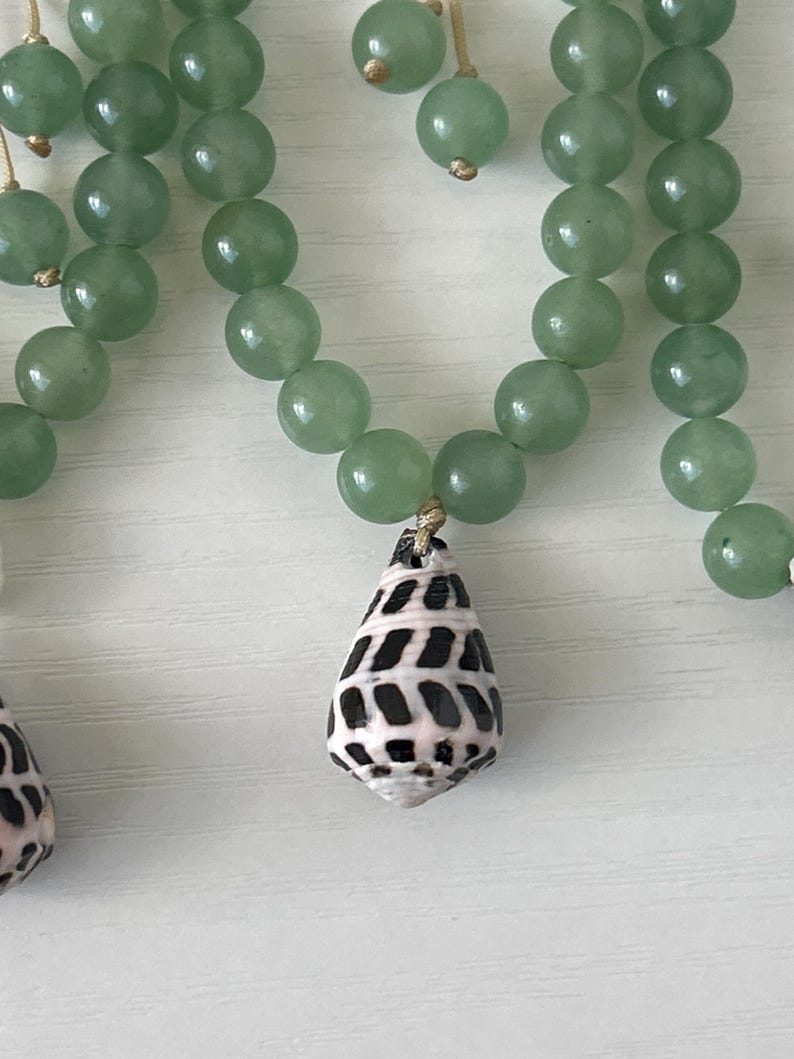 May include: A necklace featuring light green, round beads and a teardrop-shaped pendant. The pendant is a seashell with a black and white pattern. The necklace is strung on a thin, gold-colored cord.