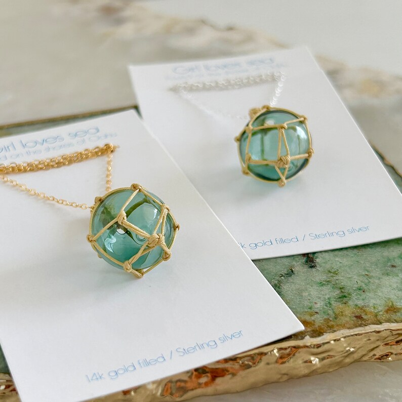 May include: Two gold necklaces with a blue glass orb pendant encased in a gold wire cage. The necklaces are displayed on white cards with the text "14k gold filled/Sterling silver".
