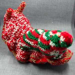 May include: A crocheted snail toy in red, white, green, and tan yarn. The snail's body is mainly red and white, with a green and red shell. It has two tan antennae and a tan base. The snail is a decorative item.