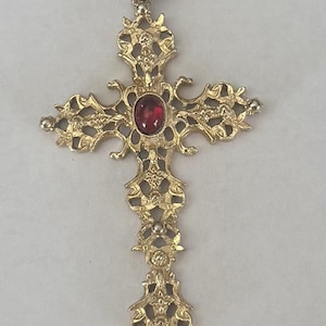 May include: A gold-toned metal cross pendant featuring an intricate, openwork filigree design with floral-like scrollwork. A single, smooth, oval-shaped deep red gemstone is set prominently in the center of the cross, which includes a top bail.