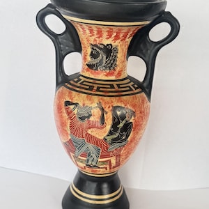 May include: A black and terracotta-colored ceramic amphora vase featuring two handles and a wide rim. The body is decorated with black-figure style illustrations of figures in profile, a Greek key pattern, and a portrait on the neck.