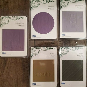 May include: Set of six embossing dies for crafting. The dies feature various designs, including a circle with a diamond pattern, a square with the text "thinking of you", a square with the text "get well soon", a square with the text "thanks", and a square with a diagonal stripe pattern. The dies are packaged in clear plastic sleeves with white backing cards.
