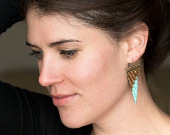 May include: Geometric earrings with a triangle design. The earrings feature a combination of brown and turquoise seed beads, creating a striking contrast. The earrings are suspended from gold-colored hooks.