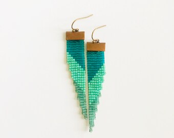 May include: A pair of handmade beaded earrings in shades of teal and green. The earrings feature a geometric design with a copper-colored top and gold-colored ear wires. The beaded fringe hangs in a zig-zag pattern.