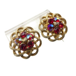 May include: A pair of vintage gold-tone clip-on earrings. Each earring features a woven, floral-like design with a cluster of red and iridescent rhinestones. The earrings are displayed on a clear, angled stand against a white background.