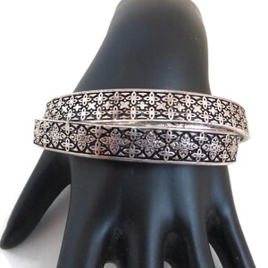 May include: Two silver-colored bangles with a floral pattern. The bangles are stacked on top of each other, and the floral design is repeated around the entire circumference of each bangle. The bangles are displayed on a black surface.