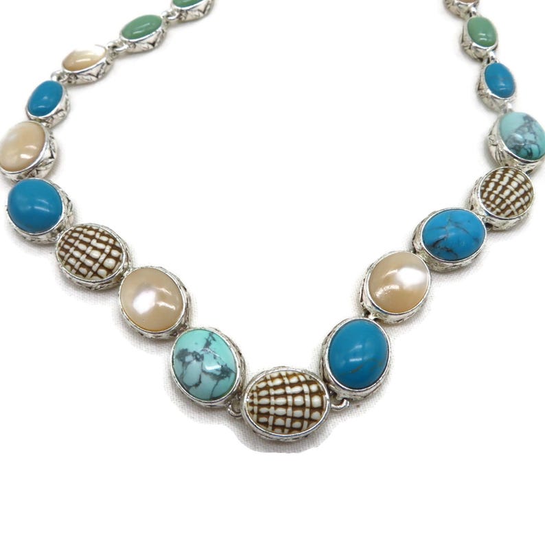 A silver-tone necklace with alternating oval and round beads in turquoise, cream, and brown, arranged in a curve against a white background.