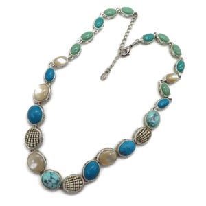 A silver-tone necklace with alternating oval and round beads in turquoise, cream, and brown, laid flat in a full circle with an extender chain visible on a white background.