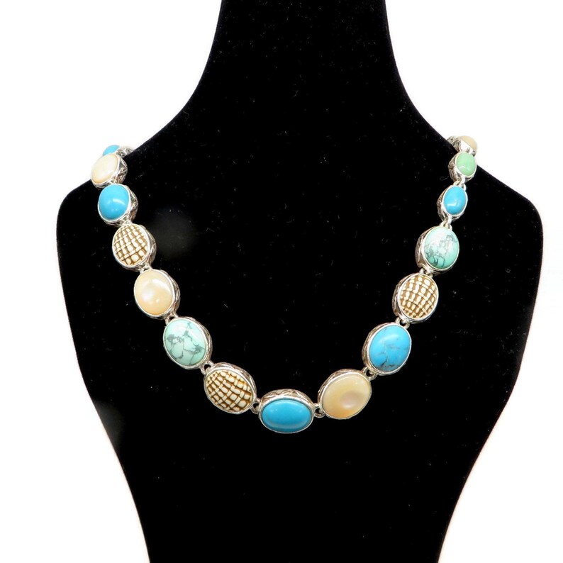 A silver-tone necklace with alternating oval and round beads in turquoise, cream, and brown, displayed on a black mannequin bust.