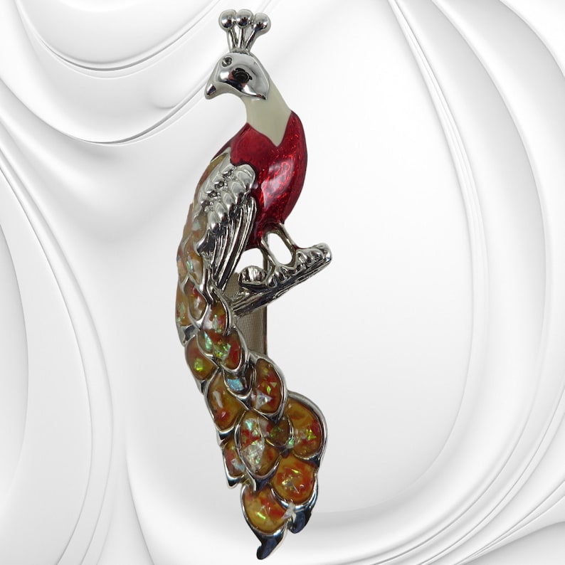 K&ouml;nnte beinhalten: Ein dekorativer Metallclip in Pfauform mit silbernem Finish. Der Pfau hat eine rote Brust, schillernde Schwanzfedern und eine Krone. Der Clip dient zum Halten von Gegenst&auml;nden und verleiht einen Hauch von Eleganz.