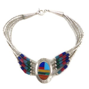 May include: A multi-strand silver necklace featuring a central oval pendant inlaid with geometric segments of blue, red, yellow, and green stone. The necklace includes silver beads and cylindrical segments of blue, red, and green stone accents.