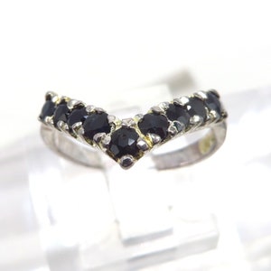 May include: A silver-toned ring with a V-shaped design, set with seven round, black gemstones. The ring is displayed on a clear surface, showcasing the contrast between the dark stones and the bright metal.