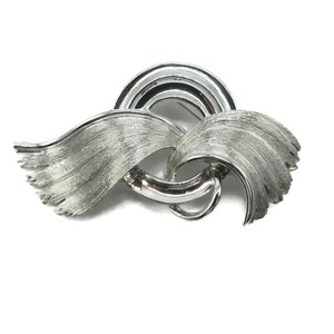 May include: Silver-toned brooch with a textured, brushed metal finish. The brooch features a curved design with a central loop and a semi-circular element at the top. The overall shape resembles stylized wings or flowing ribbons.