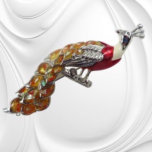 K&ouml;nnte beinhalten: Dekorative Pfauenbrosche mit silberfarbenem Metallrahmen. Die Brosche hat einen roten, wei&szlig;en und silbernen Kopf und K&ouml;rper mit schillernden orangefarbenen und gelben Schwanzfedern. Ein Accessoire f&uuml;r Kleidung.