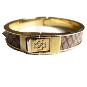 May include: Gold-tone metal hinged bracelet with a brown, textured band. The bracelet features a decorative clasp with an embossed design. The band has a snakeskin pattern. The bracelet is designed to be worn around the wrist.