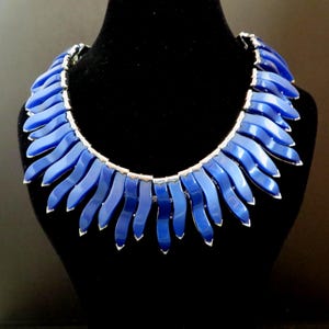 May include: A statement necklace featuring multiple curved, vibrant blue segments arranged in a fringe-like pattern. Each individual blue piece is capped with a metallic silver accent, creating a layered, textured look around the collar.