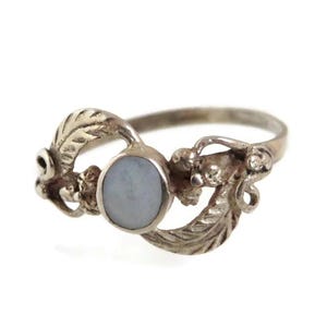 May include: A silver-toned ring featuring a central oval-shaped light blue stone. The band is decorated with sculpted leaf motifs and small spherical accents that extend from the sides of the stone setting, creating a continuous metal design.