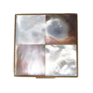 May include: A square compact case featuring a four-panel inlay of iridescent mother-of-pearl tiles. Each tile displays unique swirling patterns in shades of white, gray, and soft pink. The entire square piece is framed by a thin, textured gold-tone metal border.