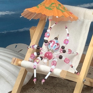 May include: A pink and white beaded spider figurine sitting on a miniature beach chair with a small orange and green umbrella.