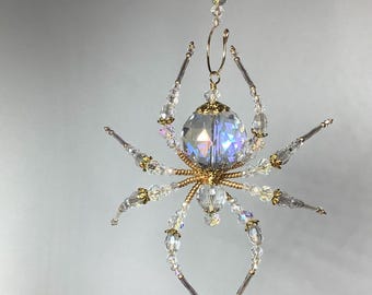 May include: A crystal spider ornament with a gold-coloured metal frame. The spider has a large, faceted crystal body and crystal legs. The ornament is designed to hang, with a loop at the top. The crystals catch the light, creating a shimmering effect.