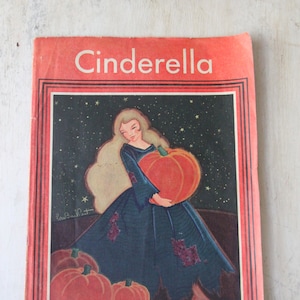 May include: A vintage children's book cover featuring Cinderella in a blue dress holding a large orange pumpkin. The cover is red with a black border and the title "Cinderella" in white letters.