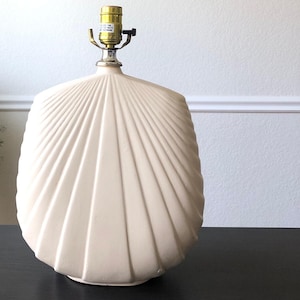 May include: A white ceramic table lamp with a fan-shaped base. The lamp has a gold-colored metal base and a white fabric shade.