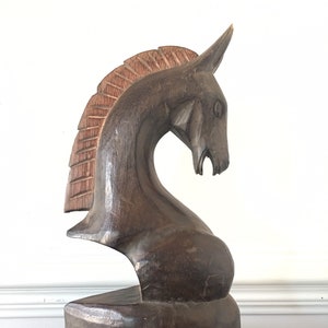 May include: A dark brown wooden horse head sculpture with a carved mane. The sculpture is sitting on top of a stack of three vintage books.