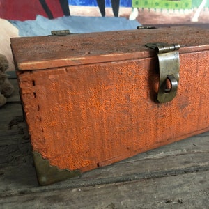 May include: A small wooden box with a hinged lid and a metal latch. The box is painted a bright orange color and has decorative metal corners.