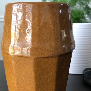May include: A brown ceramic vase with a geometric, octagonal shape. The vase has a glossy finish and is perfect for displaying flowers or greenery.