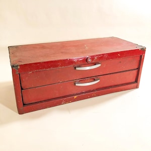 May include: A vintage red metal toolbox with two drawers and a lock. The toolbox has silver handles and shows signs of wear, with some paint chipping. The overall design is rectangular and sturdy, suggesting it's built for durability.
