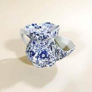 May include: A white ceramic shaving scuttle with a blue floral pattern. The scuttle has a handle, a top bowl, and a side compartment. The blue and white design covers the entire surface of the scuttle, creating a vintage aesthetic.