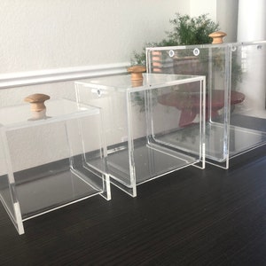 May include: Four clear acrylic boxes with wooden knobs on top, arranged in descending size order. The boxes are empty and appear to be made of thick, sturdy acrylic.