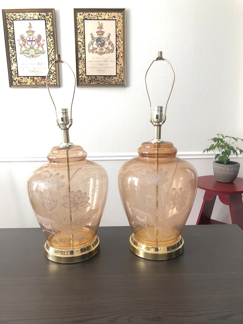 May include: Two amber glass table lamps with etched floral designs. The lamps have gold bases and are on a dark brown table.