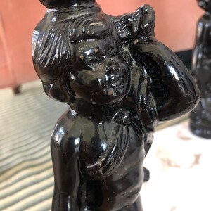May include: A black ceramic candlestick featuring a cherub figure with a detailed face and a flowing drapery. The candlestick has a smooth, glossy finish.