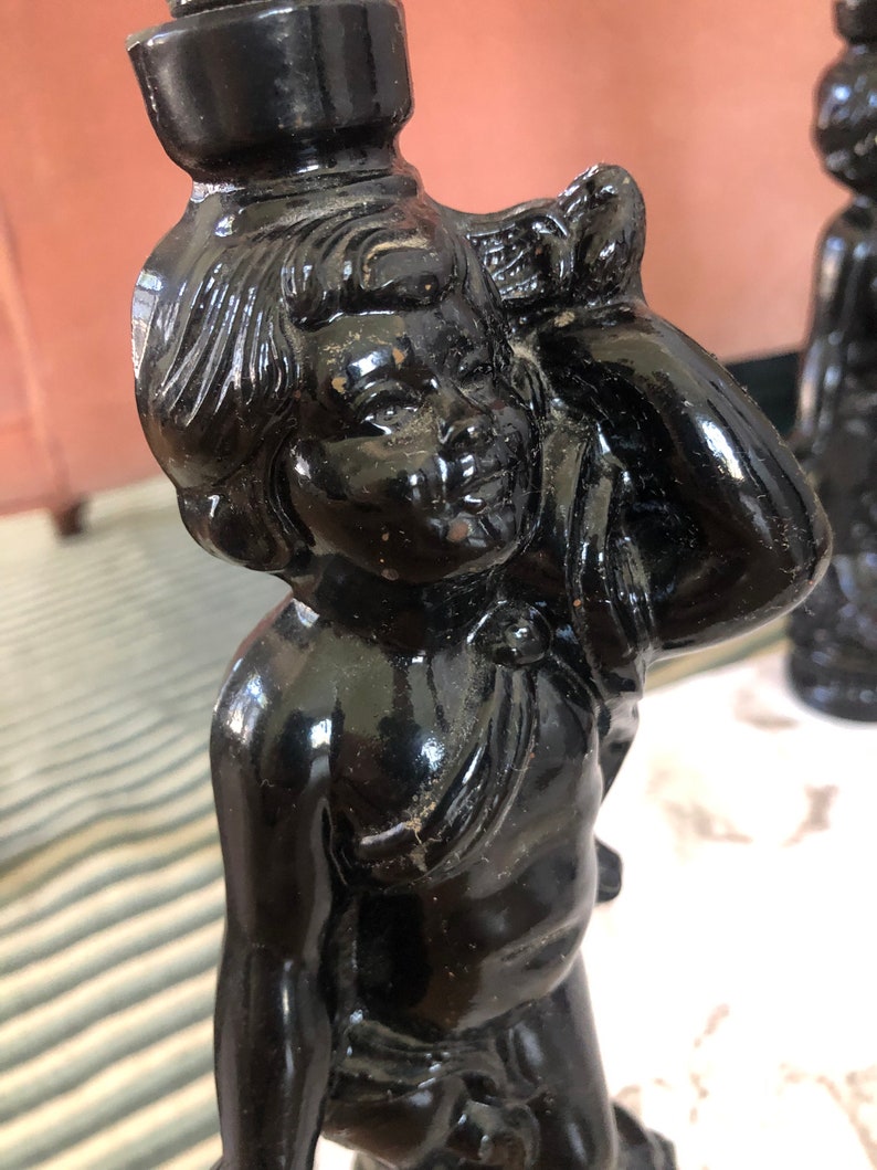 May include: A black ceramic candlestick featuring a cherub figure with a detailed face and a flowing drapery. The candlestick has a smooth, glossy finish.