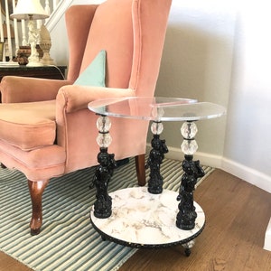 May include: A round glass-top side table with a white marble base. The table is supported by three black cherub figurines with crystal accents.
