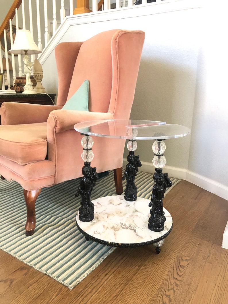 May include: A round glass-top side table with a white marble base. The table is supported by three black cherub figurines with crystal accents.