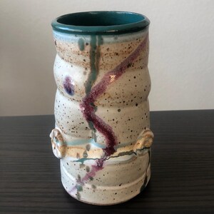 May include: A white ceramic vase with a teal rim and brown, purple, and blue glaze. The vase has a textured surface and a unique, hand-crafted design.