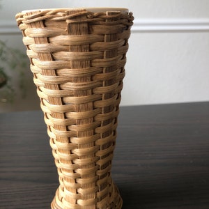 May include: A woven wicker vase with a white ceramic base. The vase is tall and narrow, with a cylindrical shape. The wicker is a light brown color, and the ceramic base is white.