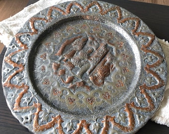 May include: A round silver and copper tray with an intricate design. The center of the tray features a raised design of figures in a stylized floral pattern. The tray has a copper border with a repeating pattern.