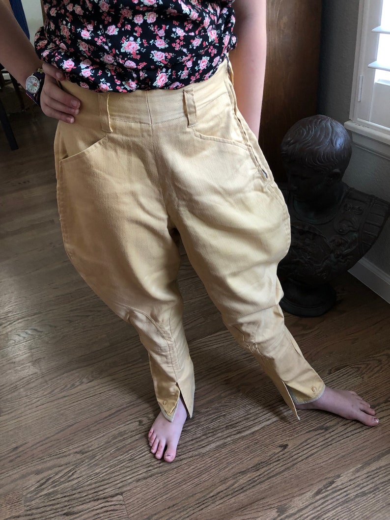 Op de afbeelding: Een beige rijbroek met een split aan de onderkant van elk been. De broek heeft een hoge taille en twee zakken aan de voorkant.