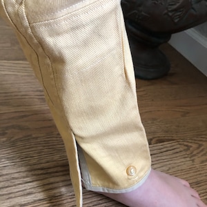 Op de afbeelding: Een gele broek met een witte bies en een knoop aan de onderkant van het been.